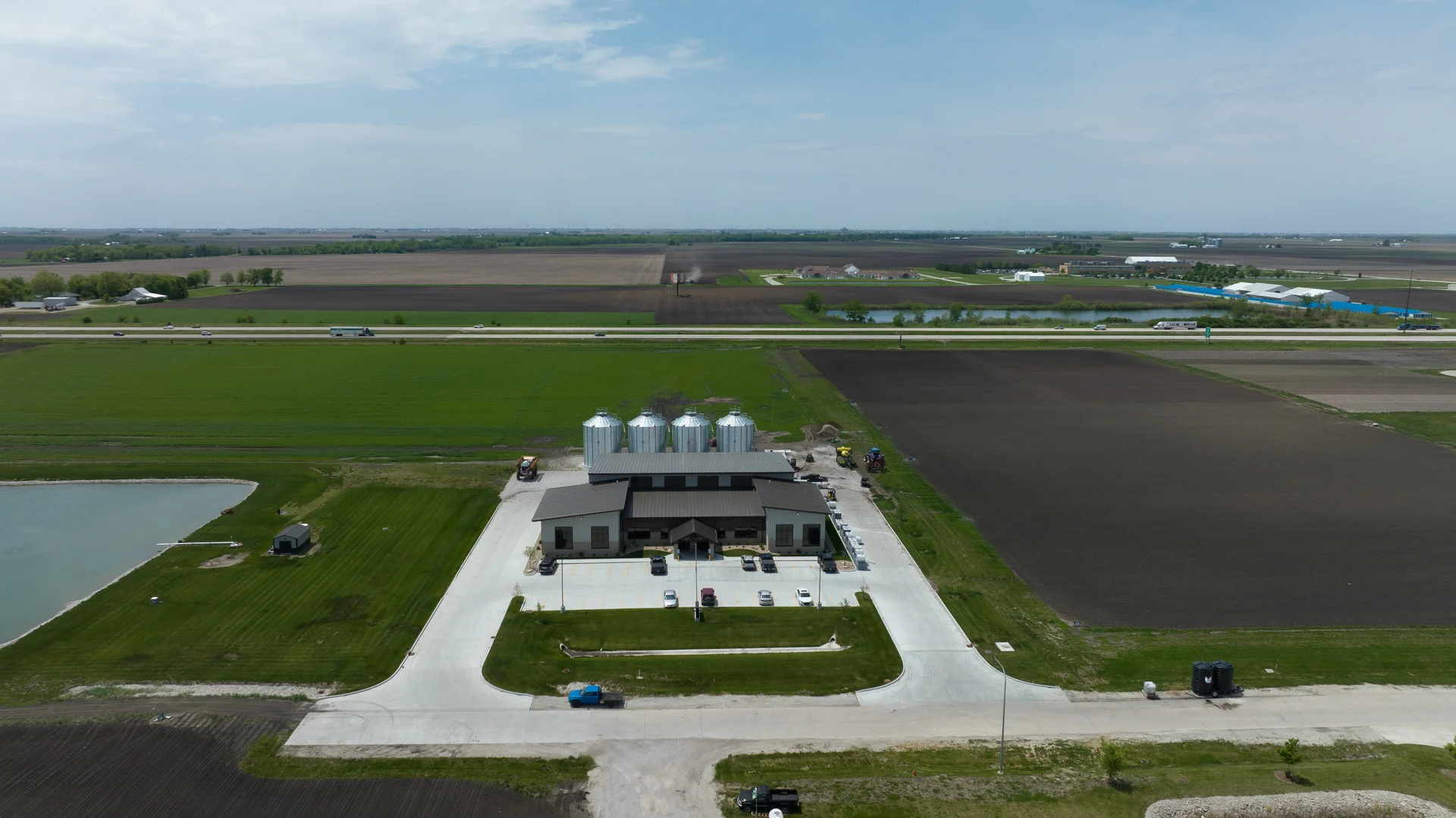 DJI_0260 Aerial view of an Agriteer precision farming facility surrounded by green fields and tilled farmland, showing a central building with parked vehicles, four grain silos behind it, concrete driveway and a nearby irrigation pond under a wide sky